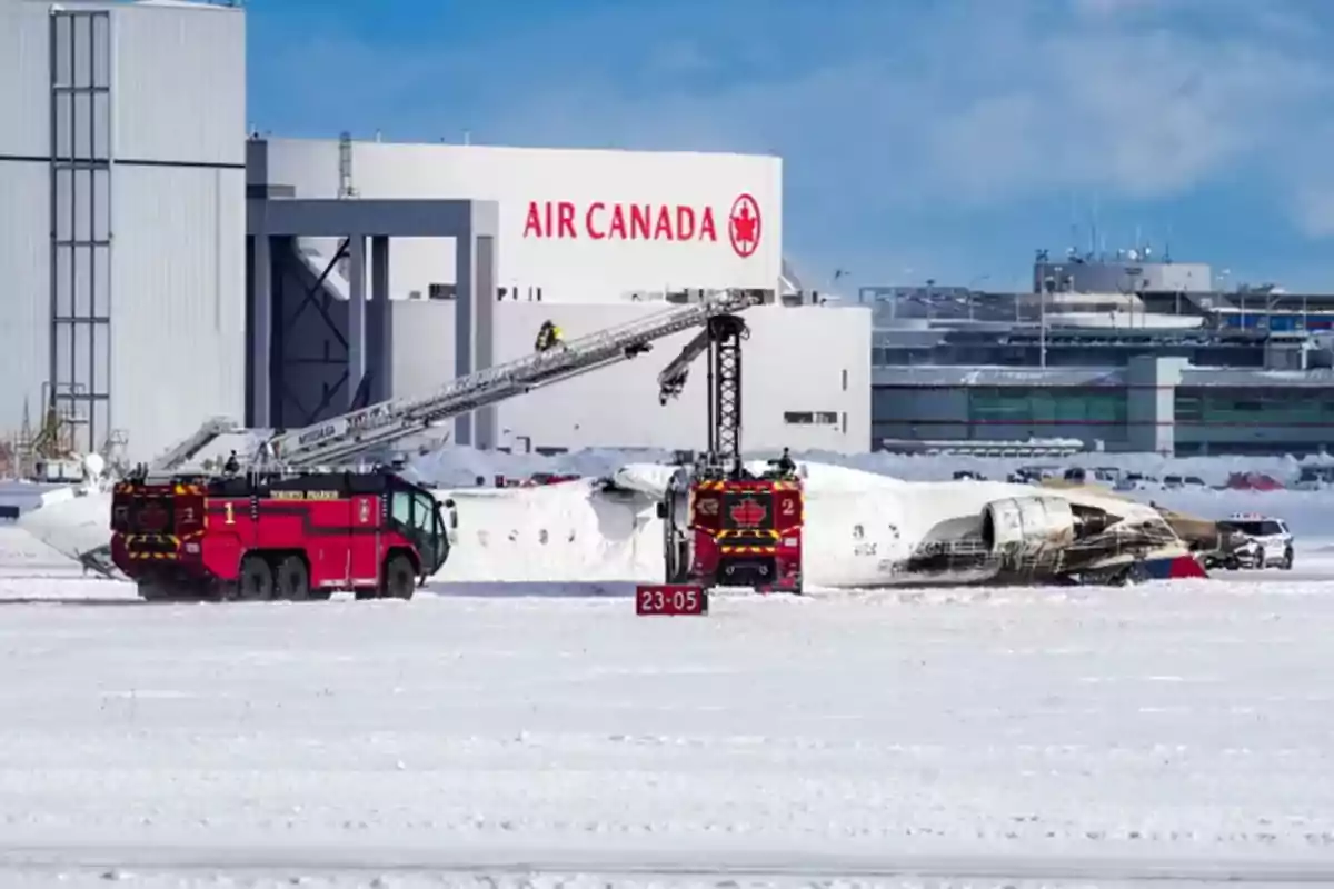 Un avión accidentado cubierto de nieve en un aeropuerto con vehículos de emergencia y un edificio de Air Canada al fondo.