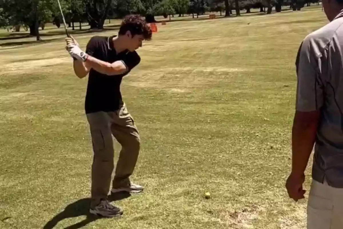 Un joven está a punto de golpear una pelota de golf en un campo mientras otra persona observa.