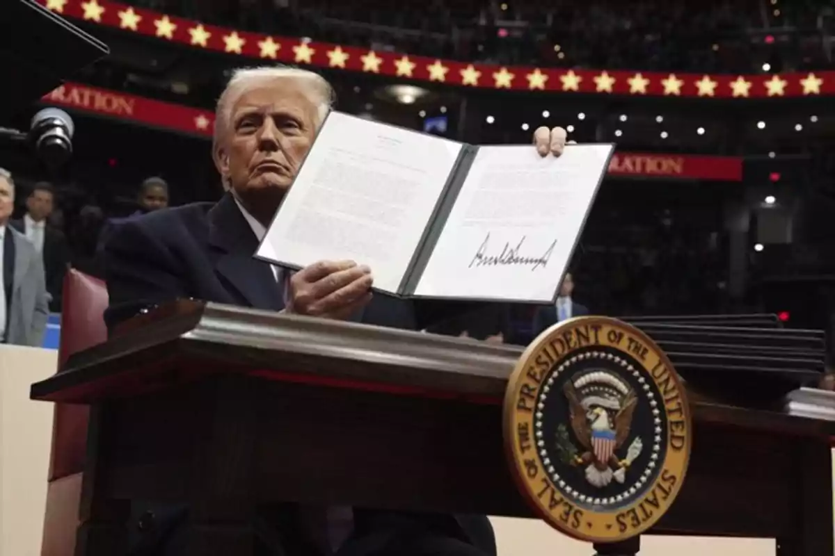 Un hombre sentado en un escritorio muestra un documento abierto con una firma visible, con un sello presidencial en el frente del escritorio y un fondo de estrellas rojas.