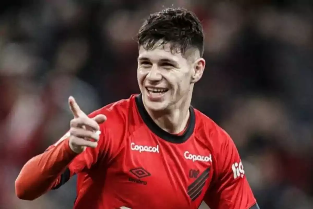 Un jugador de fútbol sonríe y señala con el dedo mientras viste una camiseta roja en un campo de juego.