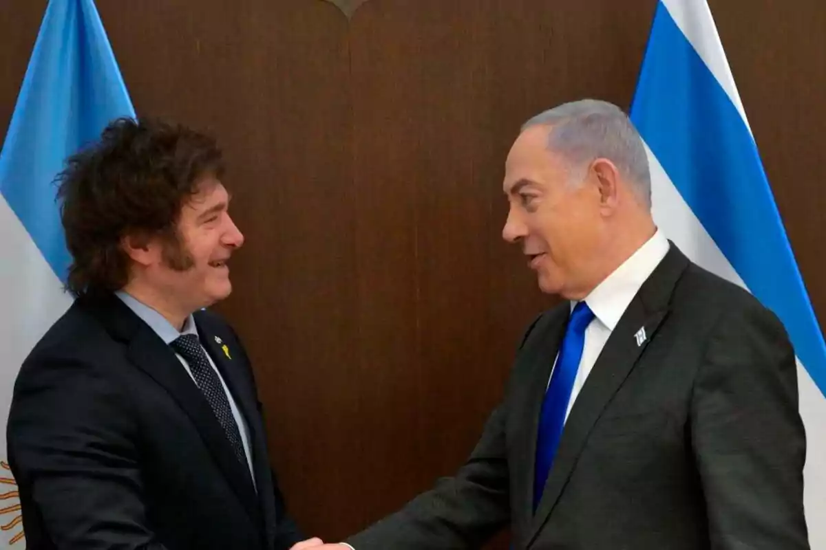 Dos hombres de traje se dan la mano frente a banderas de Argentina e Israel.