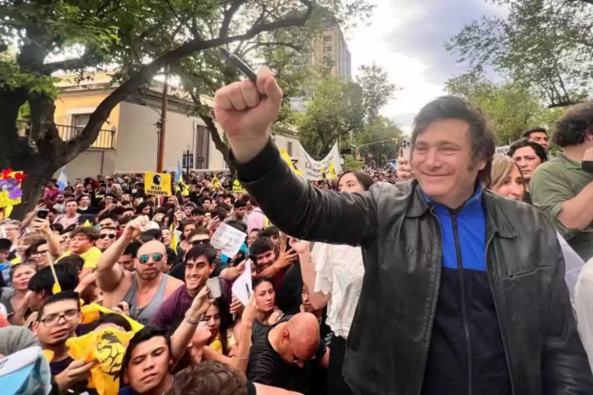 Una persona sonriente con chaqueta de cuero levanta el puño en medio de una multitud durante una manifestación al aire libre.