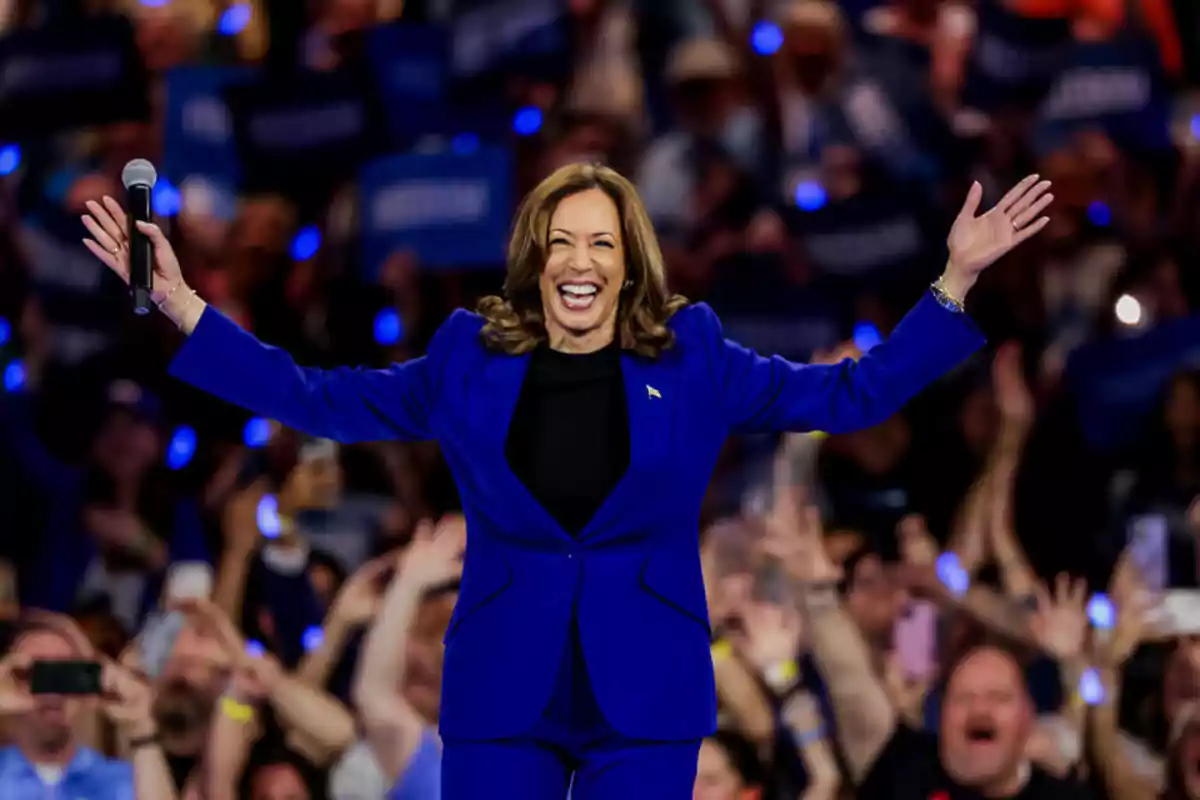 Una mujer sonriente con traje azul y los brazos extendidos, sosteniendo un micrófono, frente a una multitud animada.