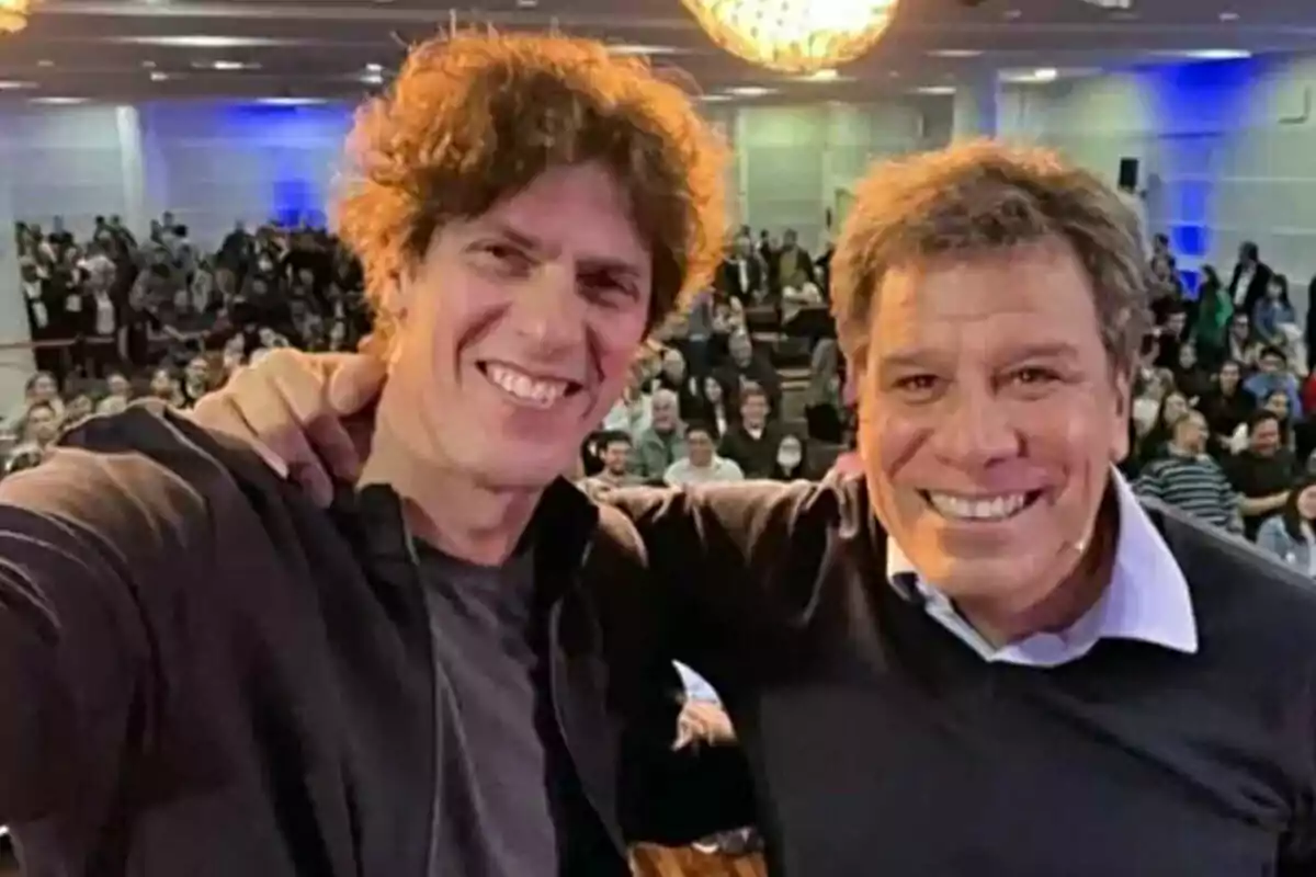 Dos personas sonrientes posan para una selfie en un evento con una multitud al fondo.