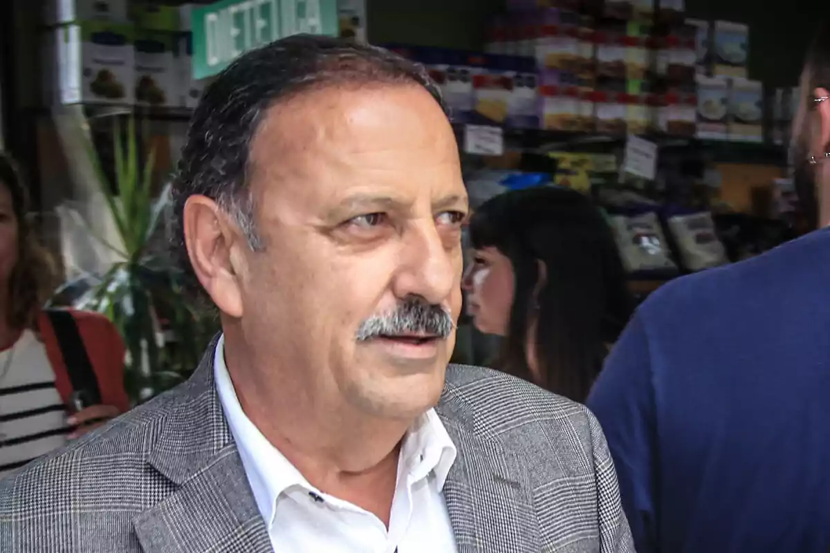 Un hombre de mediana edad con bigote y chaqueta a cuadros está en un entorno urbano con personas y productos de fondo.