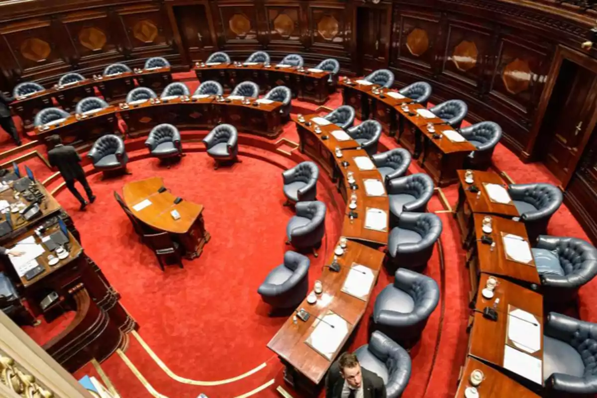 Vista de un salón parlamentario con sillas y escritorios dispuestos en semicírculo sobre una alfombra roja.