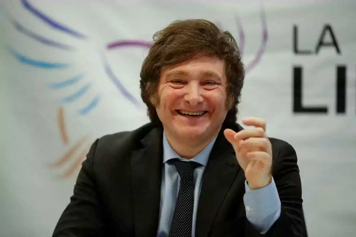 Un hombre sonriente con traje oscuro y corbata negra frente a un fondo con texto parcialmente visible.