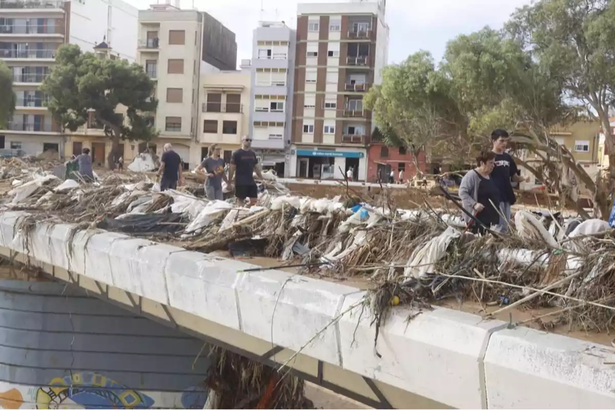 Personas caminando sobre un puente cubierto de escombros y vegetación tras una inundación en una zona urbana.