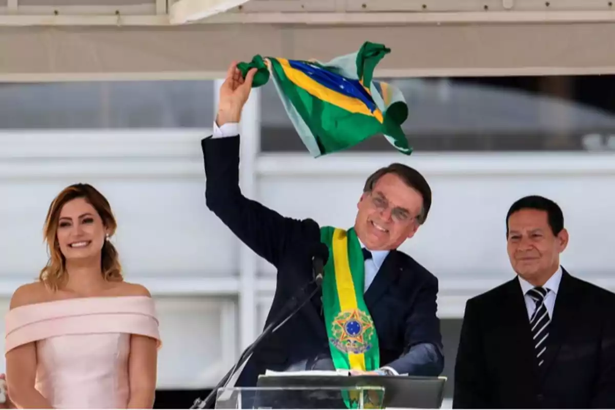 Un hombre con una banda presidencial levanta una bandera de Brasil mientras sonríe junto a una mujer y otro hombre en un evento oficial.