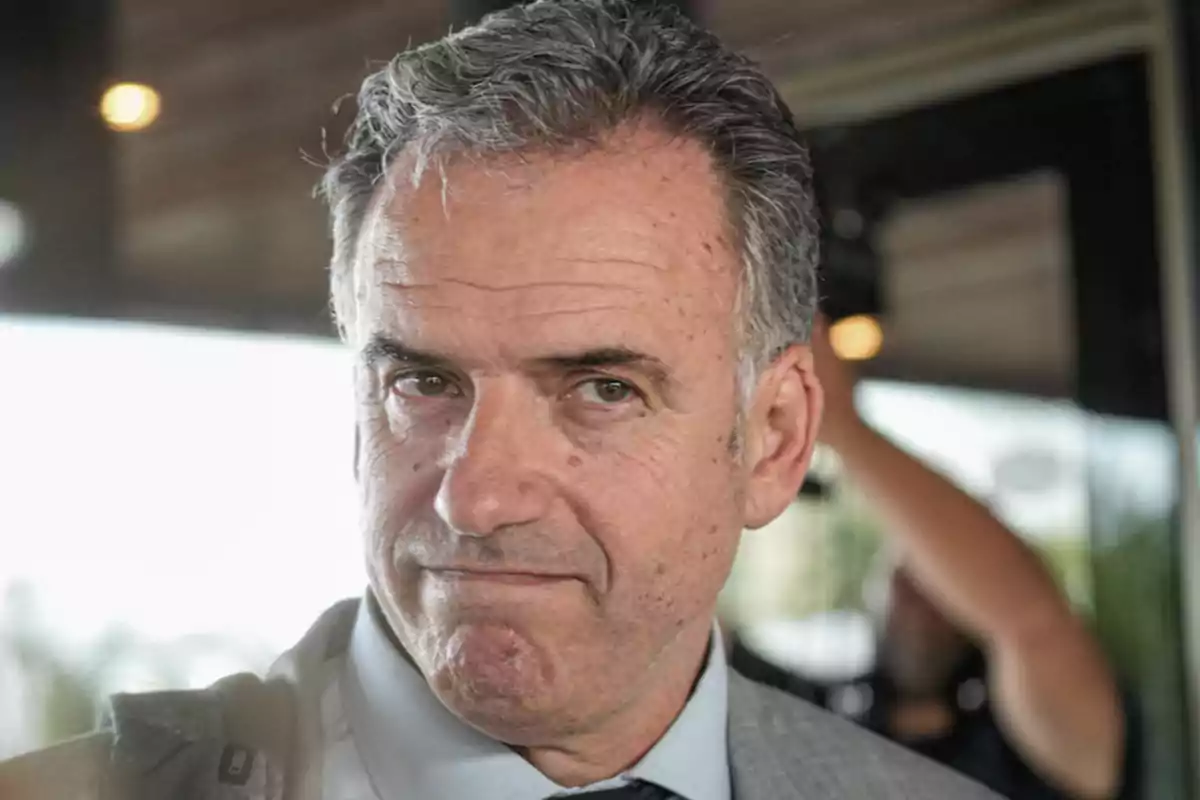 Un hombre de cabello canoso y expresión seria viste un traje gris y corbata en un entorno interior.