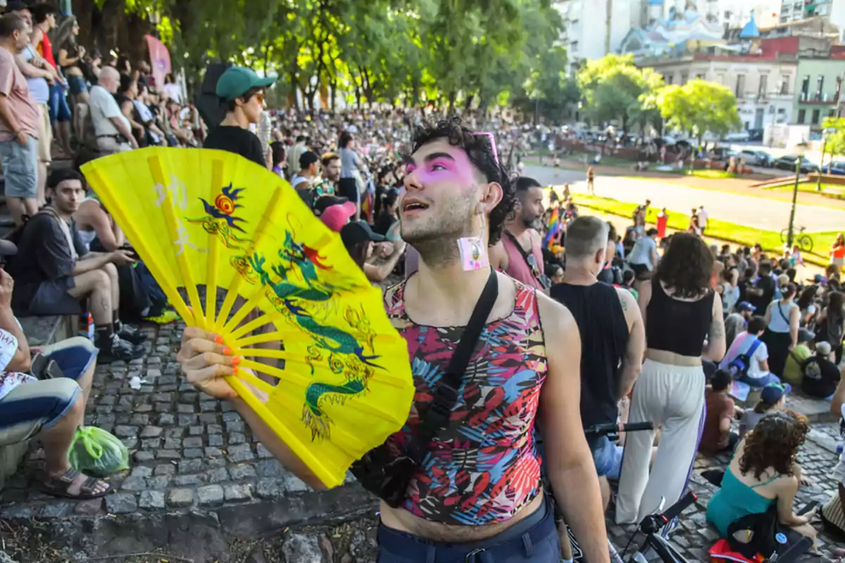 Una persona con maquillaje colorido sostiene un abanico amarillo con un diseño de dragón en un evento al aire libre lleno de gente.