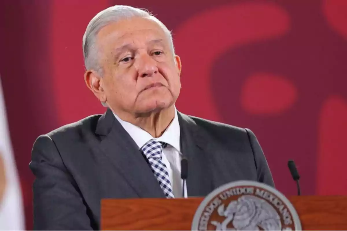 A gray-haired man in a dark suit is standing behind a podium with a microphone, against a red background.