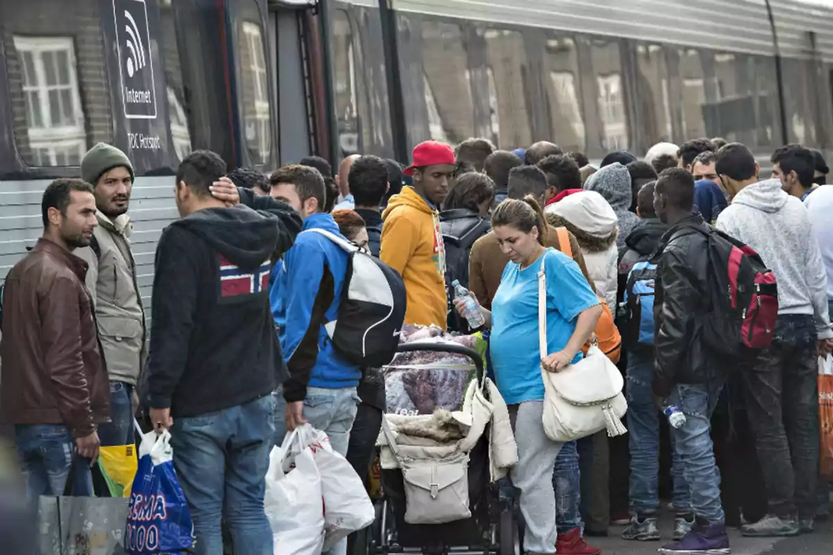 Un grupo de personas con equipaje se encuentra junto a un tren, algunas llevan mochilas y bolsas, y una mujer empuja un cochecito de bebé.