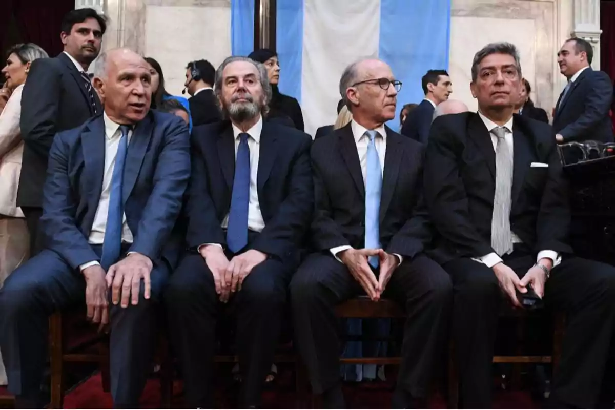 Un grupo de hombres en trajes formales está sentado en una fila durante un evento, con una bandera azul y blanca de fondo.