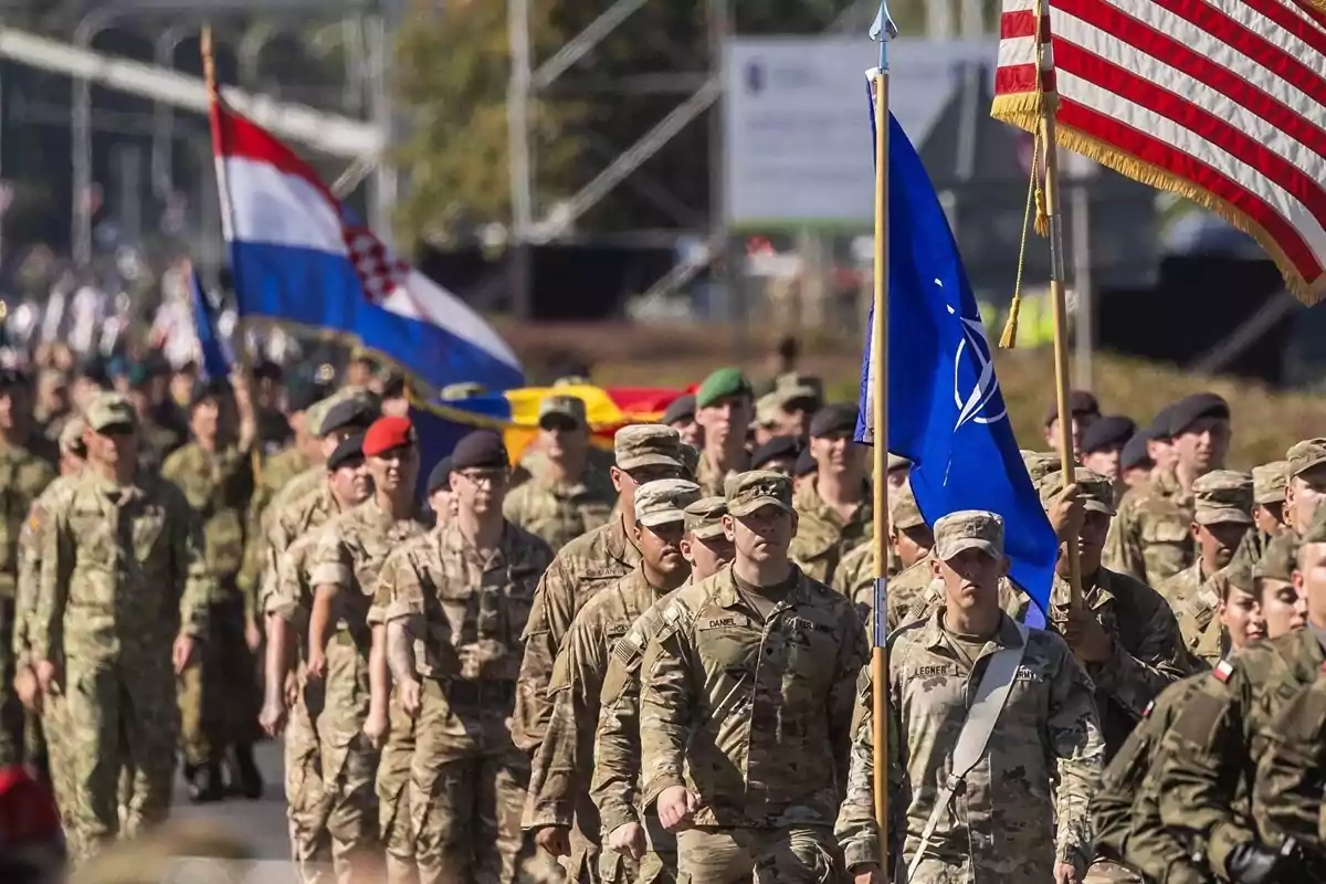 Un grupo de soldados marcha en formación, llevando banderas de varios países y de la OTAN.