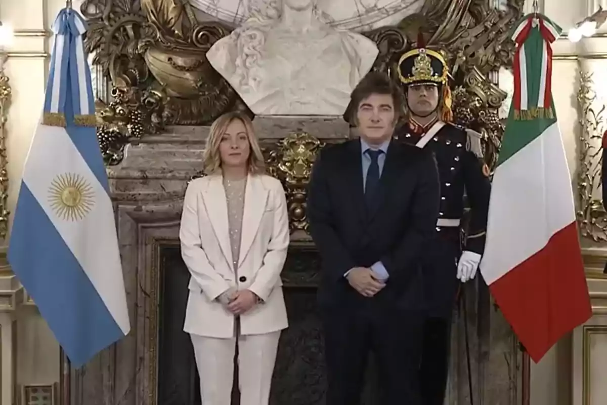 Dos personas posan frente a banderas de Argentina e Italia en un entorno elegante con un guardia detrás.