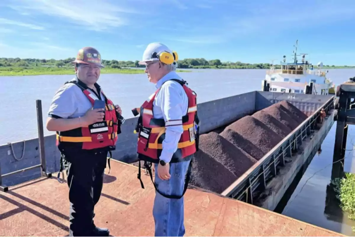 Two people wearing life jackets and safety helmets are standing next to a boat loaded with material on a river.