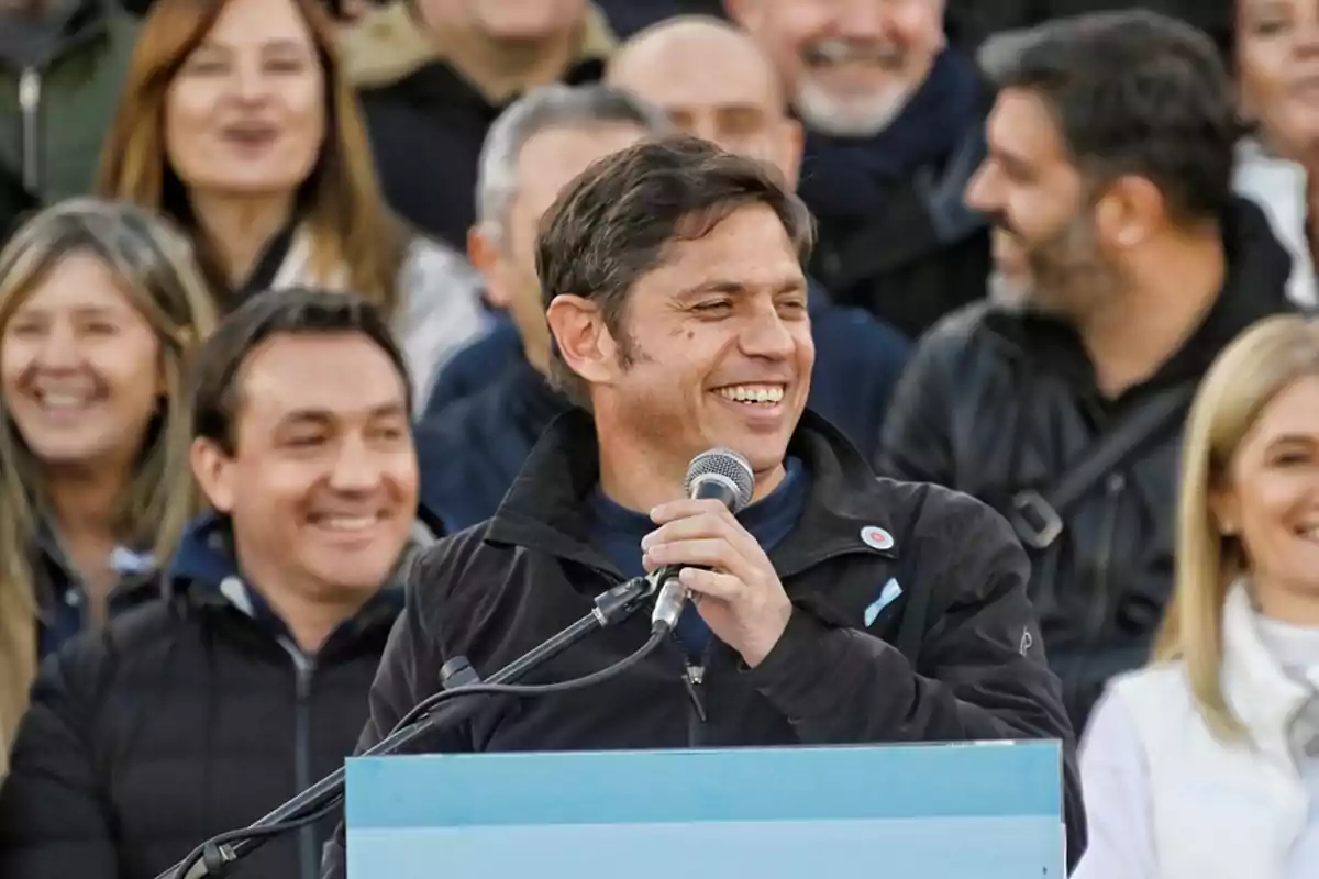 Un hombre sonriente hablando por un micrófono en un evento al aire libre rodeado de personas que también sonríen.