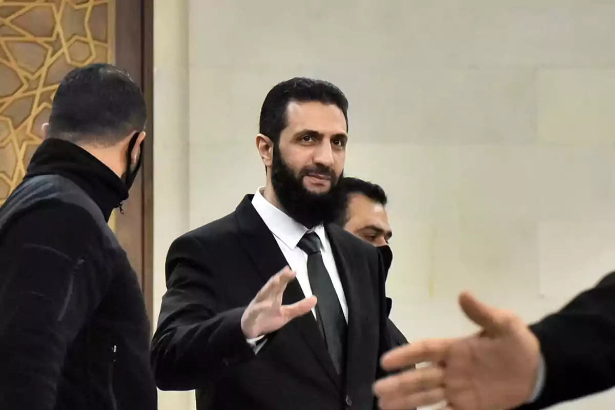 Un hombre con barba y traje oscuro está saludando mientras camina junto a otras personas en un entorno formal.