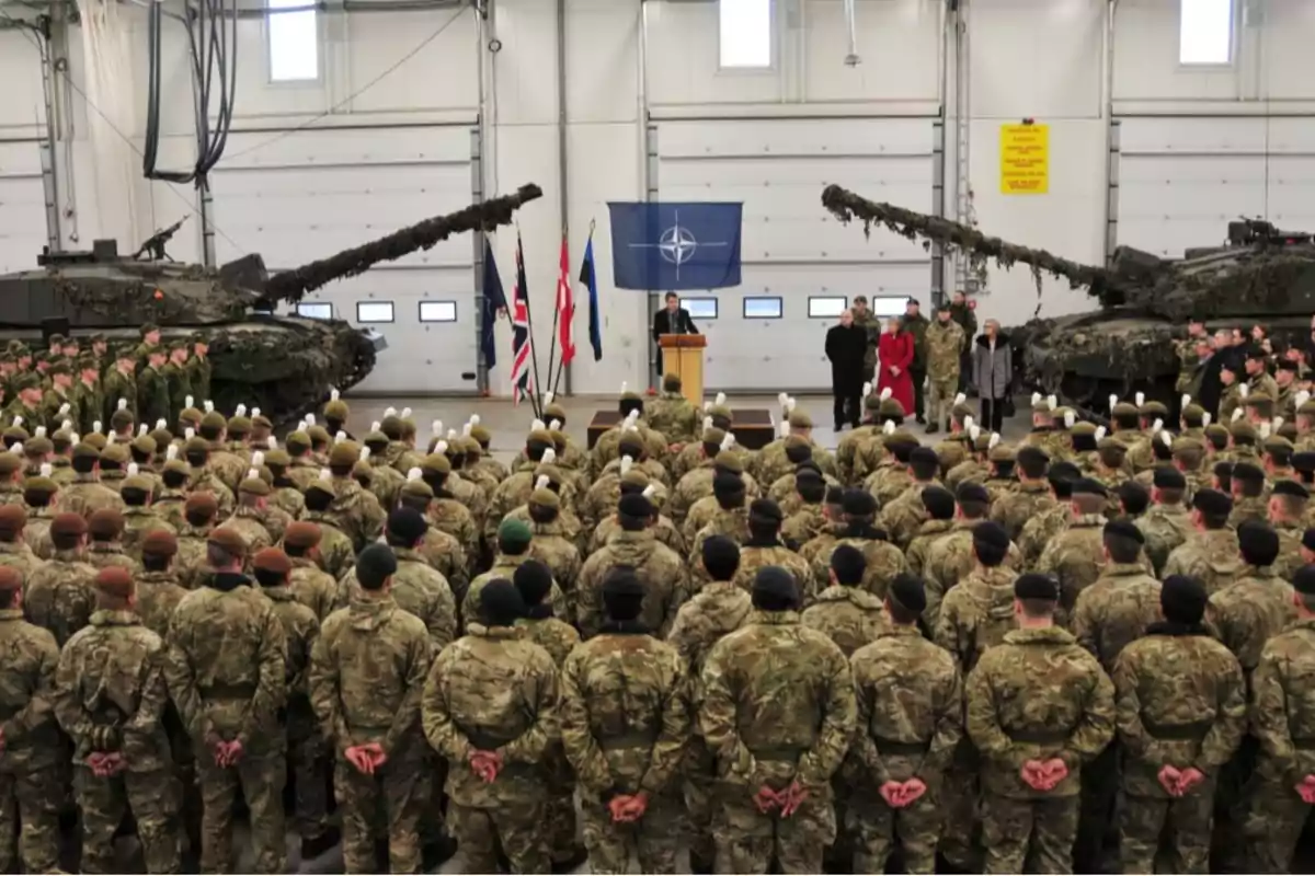 Soldados en formación escuchan un discurso en un hangar militar con tanques y banderas de la OTAN y de varios países.