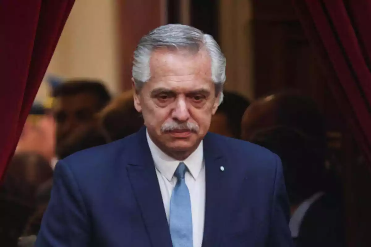 Un hombre de cabello canoso y bigote, vestido con un traje azul y corbata celeste, se encuentra en un entorno formal con cortinas rojas en el fondo.