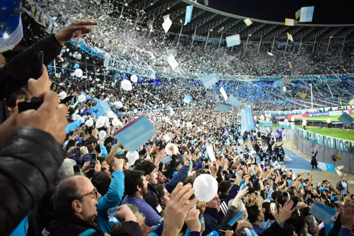 Una multitud de aficionados celebra en un estadio lanzando confeti y sosteniendo globos y pancartas mientras disfrutan de un evento deportivo.