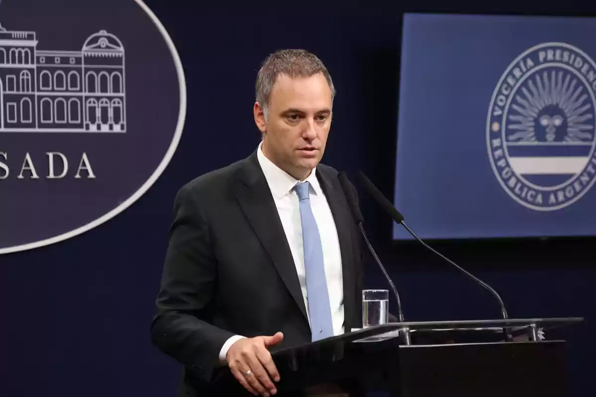 A man in a dark suit and light blue tie speaks at a podium with a microphone, with a background showing an official logo and the word "CASADA."