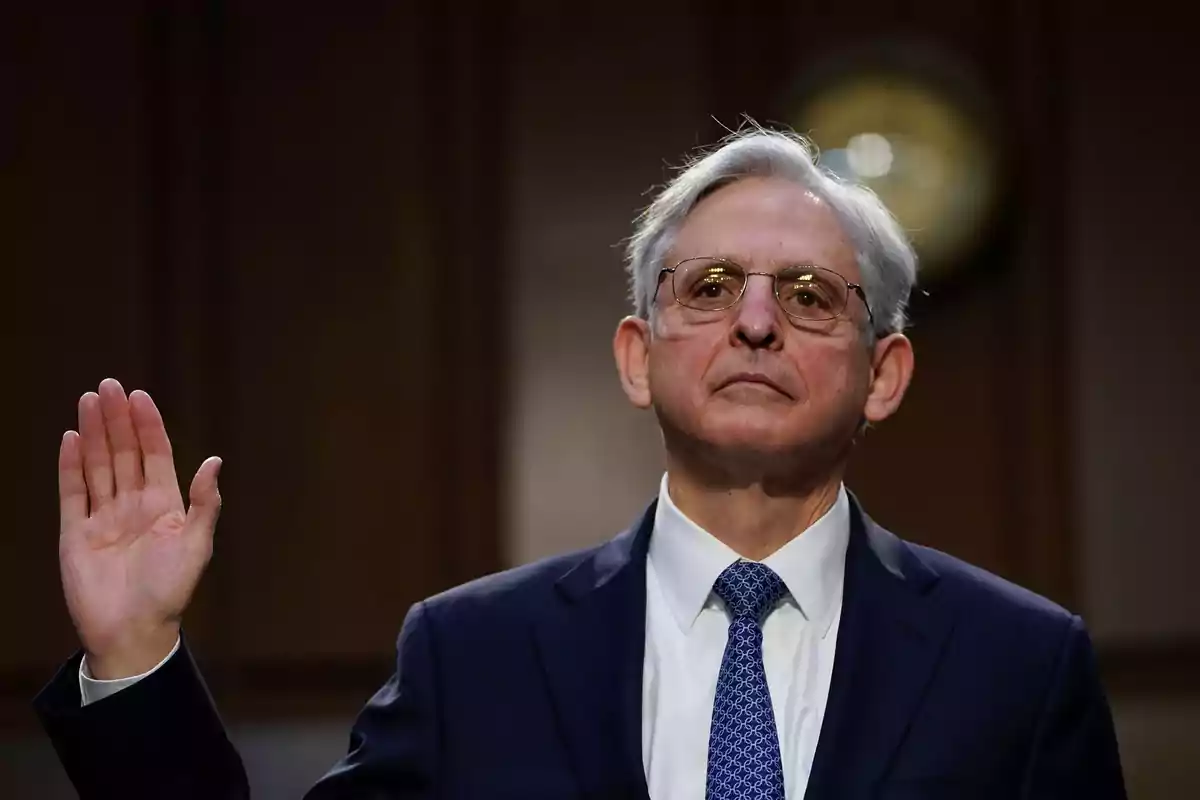 Un hombre de cabello canoso y gafas levanta la mano derecha mientras viste un traje oscuro y corbata azul en un entorno formal.