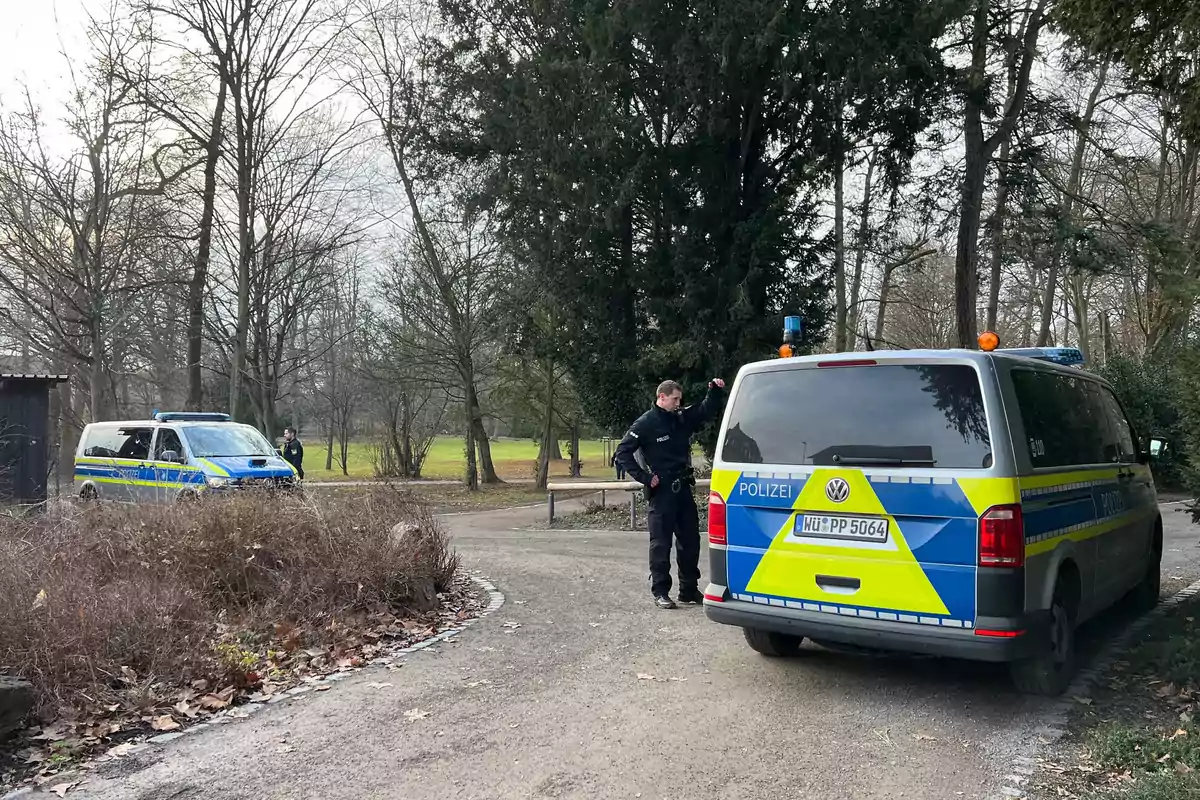 Un oficial de policía está de pie junto a una camioneta de la policía en un camino rodeado de árboles en un parque.