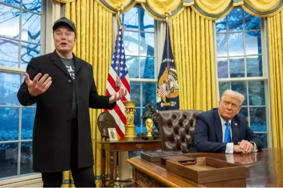 Two people in an office with golden curtains and American flags, one standing and gesturing and the other sitting behind a desk.