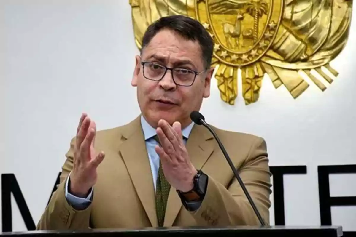 A man with glasses and a brown suit speaks into a microphone in front of a golden shield on the wall.