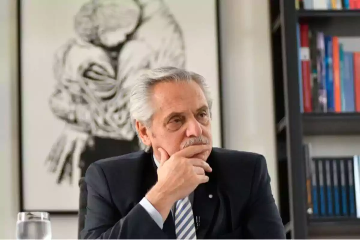 Un hombre de cabello canoso y bigote, vestido con traje y corbata, está sentado pensativo con la mano en el mentón. Detrás de él hay una obra de arte en blanco y negro que muestra a dos personas abrazándose, y a su lado hay una estantería con libros.