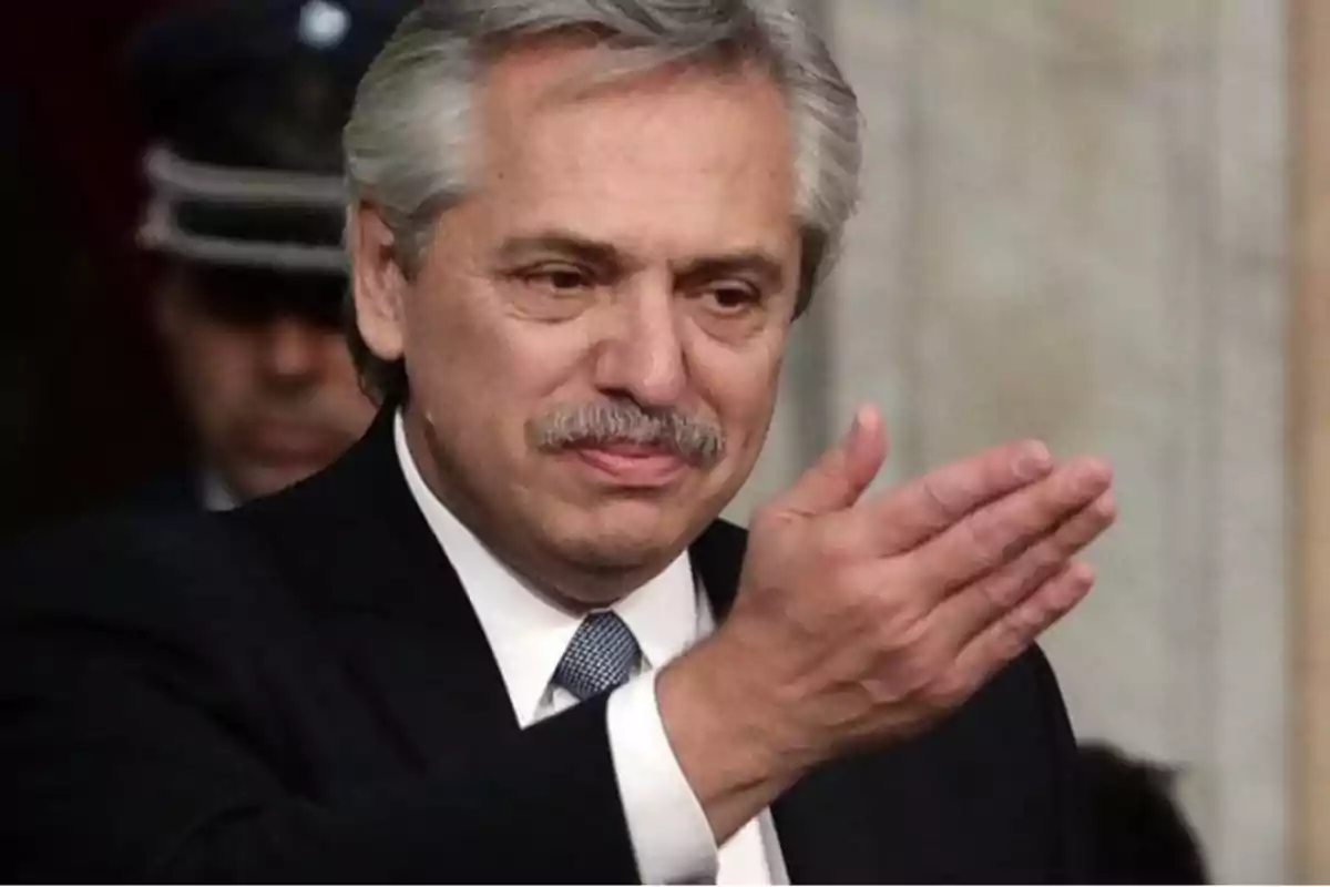 Hombre de cabello canoso y bigote, vestido con traje y corbata, haciendo un gesto con la mano.