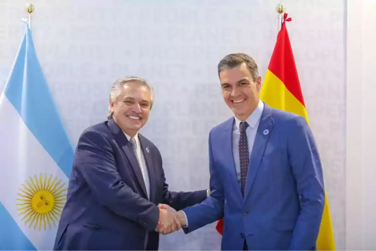 Dos hombres de traje se dan la mano frente a las banderas de Argentina y España.