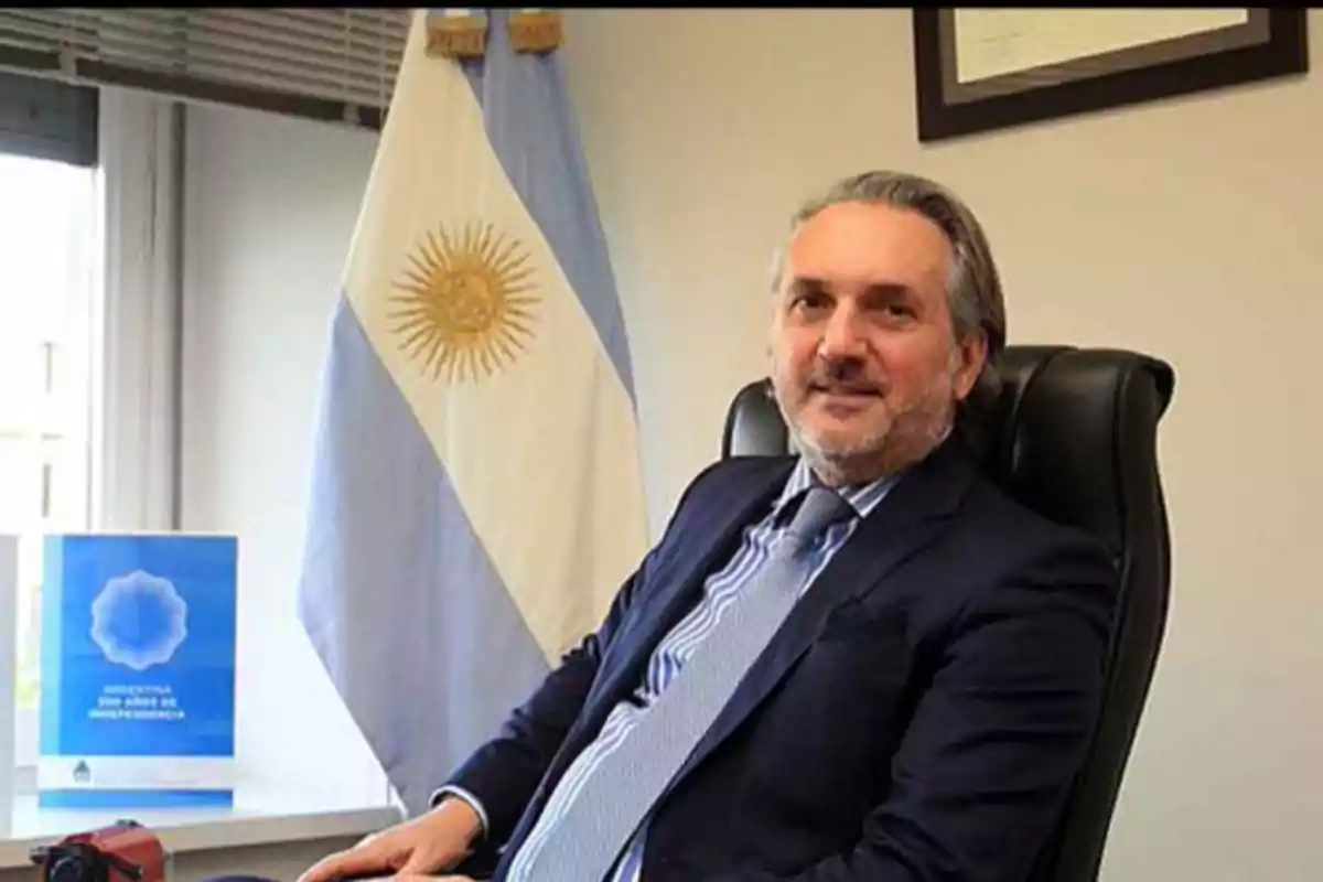 Hombre sentado en una oficina con una bandera de Argentina y un libro azul en el fondo.