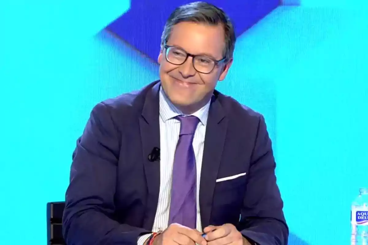 Un hombre con gafas y traje sonríe mientras está sentado en un estudio de televisión con fondo azul.