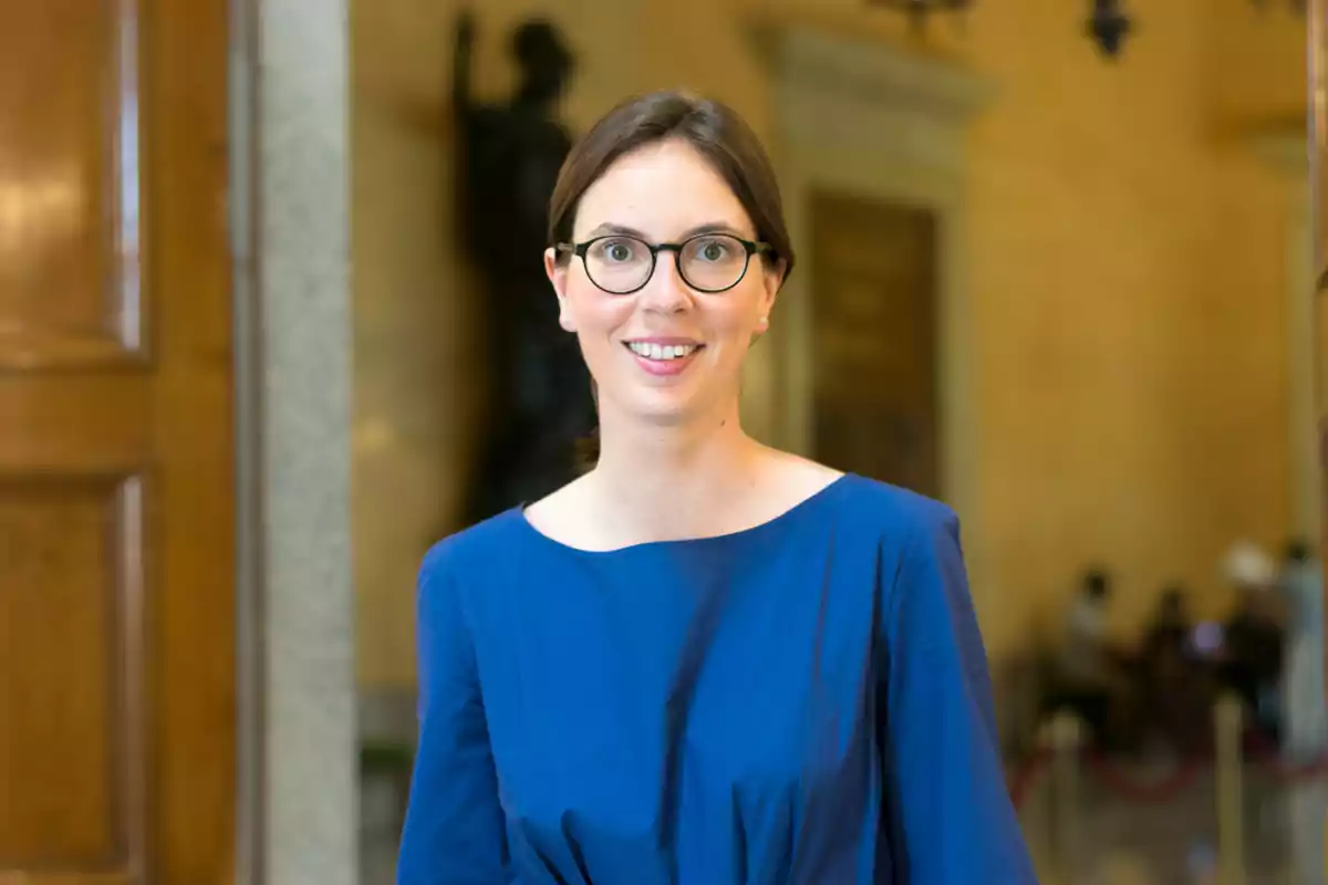 Una persona con gafas y vestido azul sonriendo en un interior elegante.