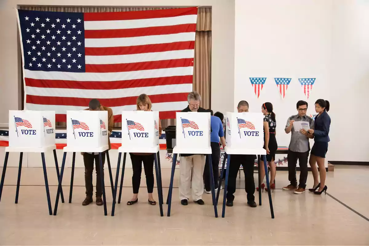 Personas votando en cabinas con una bandera de Estados Unidos al fondo.