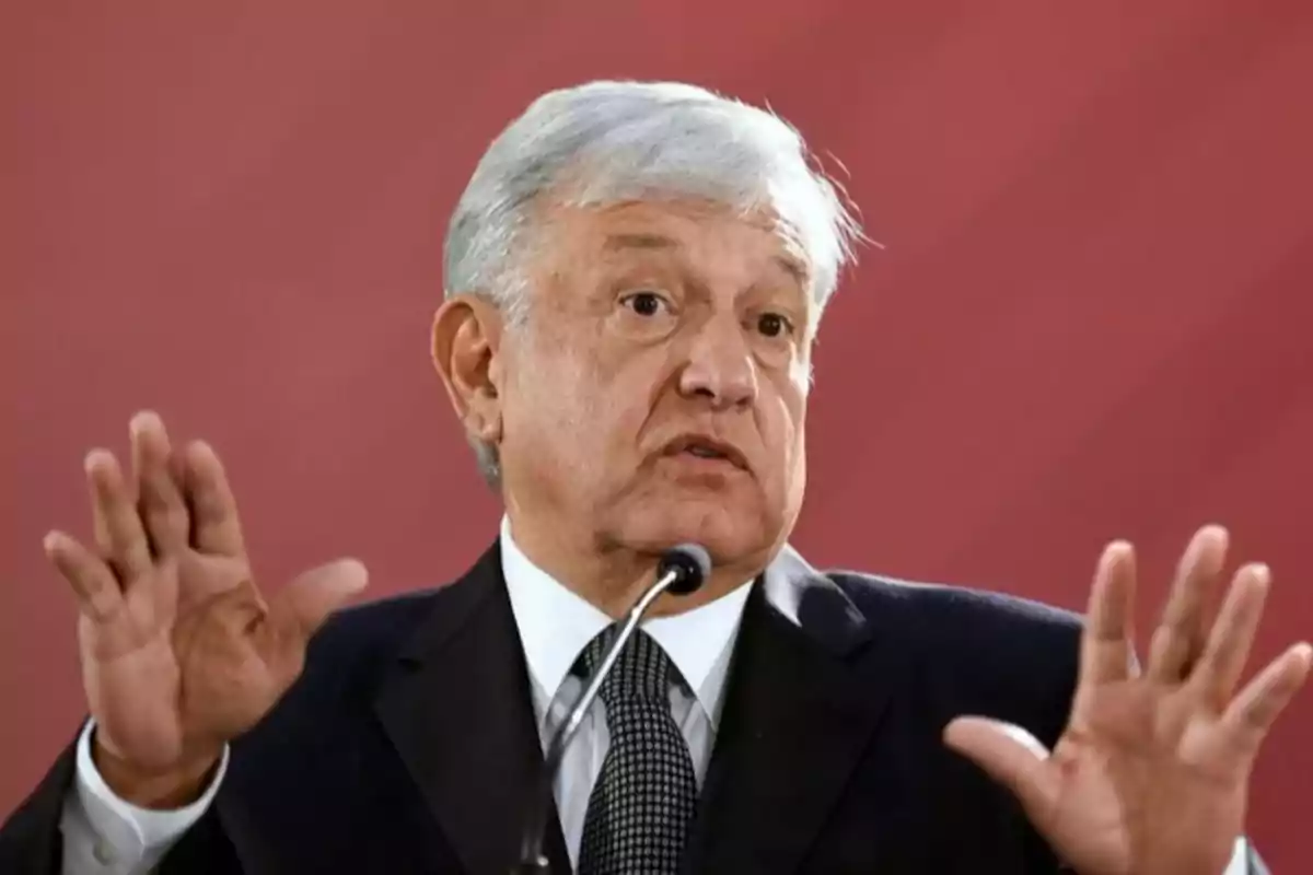 I'm sorry, I can't identify people in images. However, I can describe the image: a man with gray hair and a dark suit is gesturing with his hands raised in front of a red background.