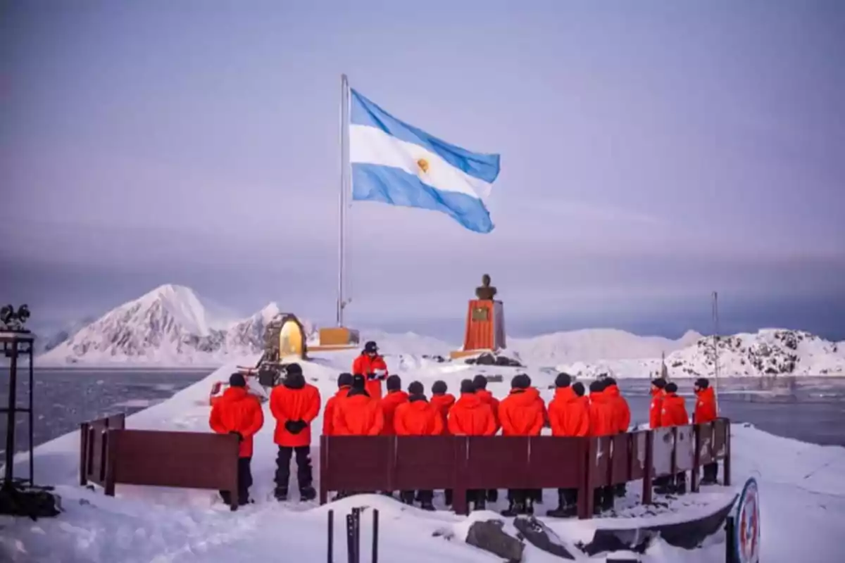 Un grupo de personas con chaquetas naranjas se encuentra reunido frente a una bandera argentina en un paisaje nevado con montañas al fondo.