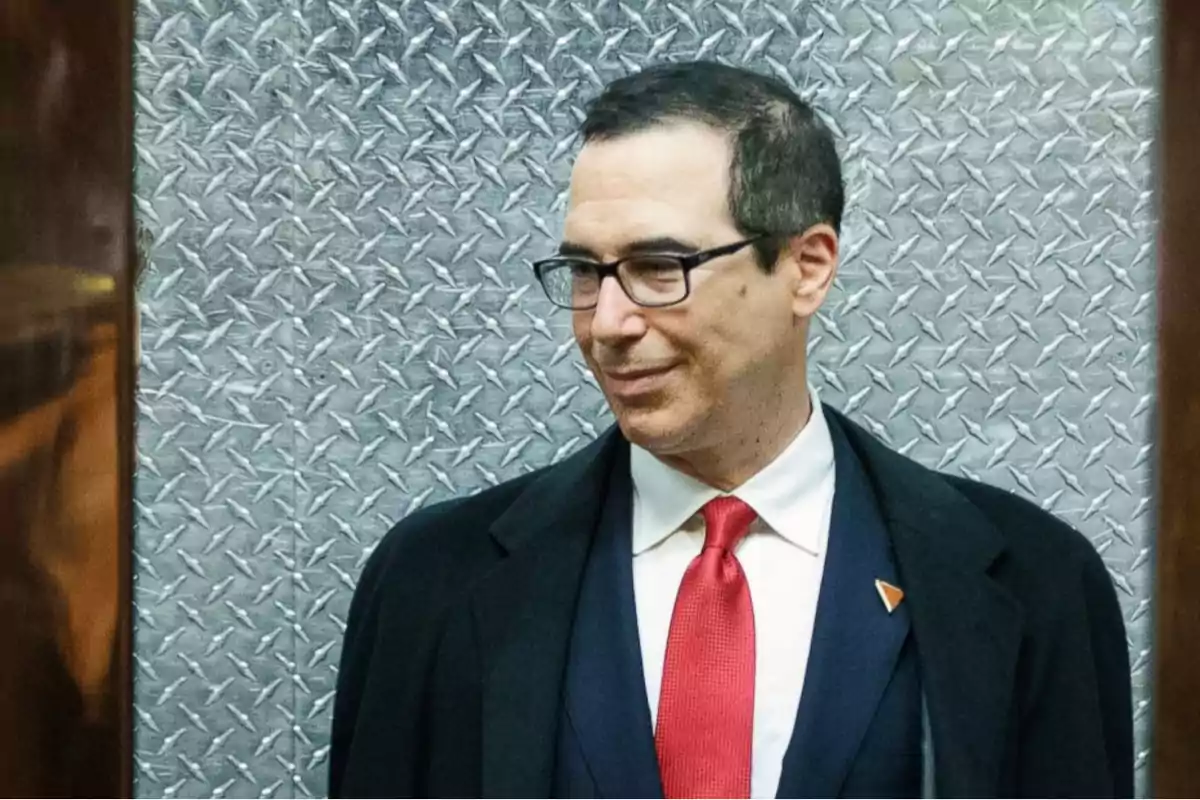 Un hombre con gafas y traje oscuro con corbata roja frente a un fondo de metal texturizado.