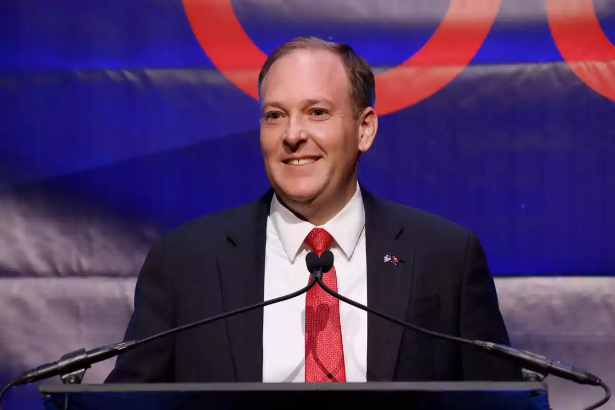 A man in a dark suit and red tie speaking at a podium with a blue and red background.