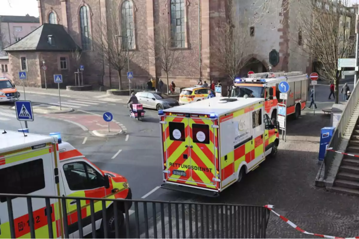 Una escena urbana con vehículos de emergencia, incluyendo ambulancias y un camión de bomberos, estacionados cerca de un edificio antiguo mientras personas caminan por la acera.