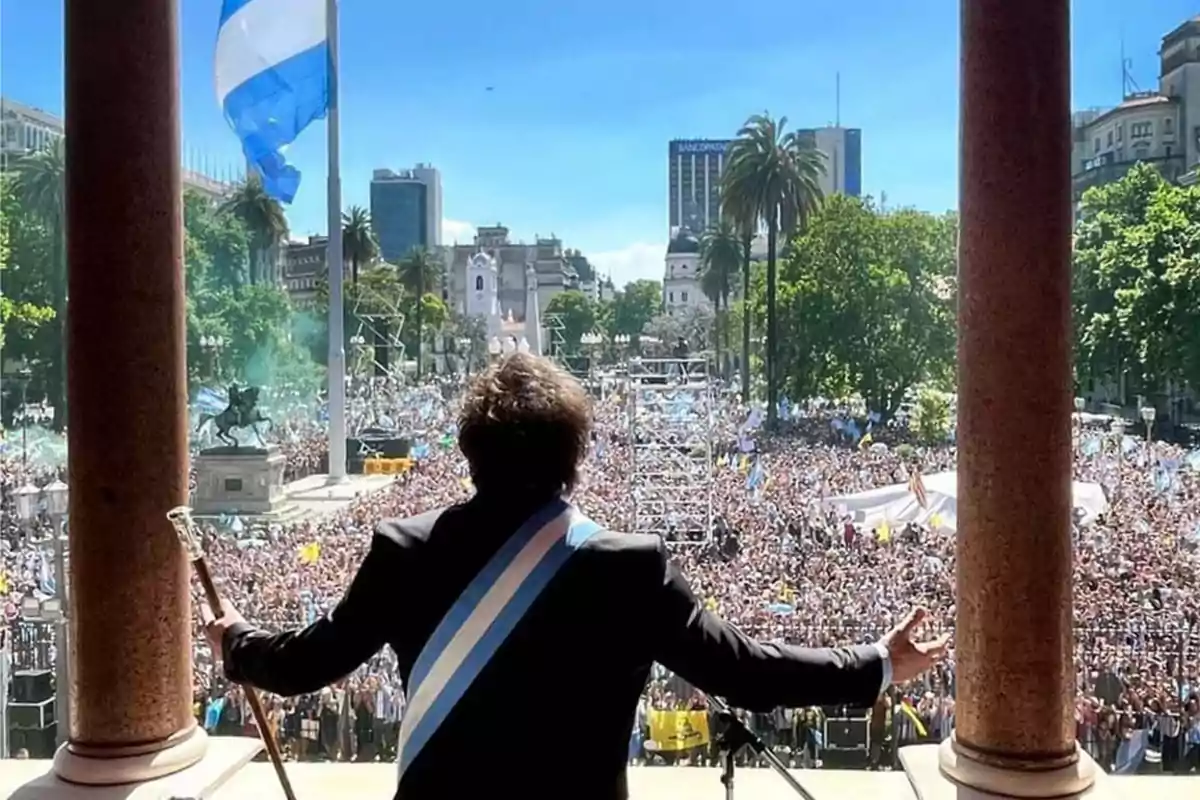 Una persona con banda presidencial y bastón de mando se dirige a una multitud desde un balcón, con una bandera ondeando y edificios de fondo.