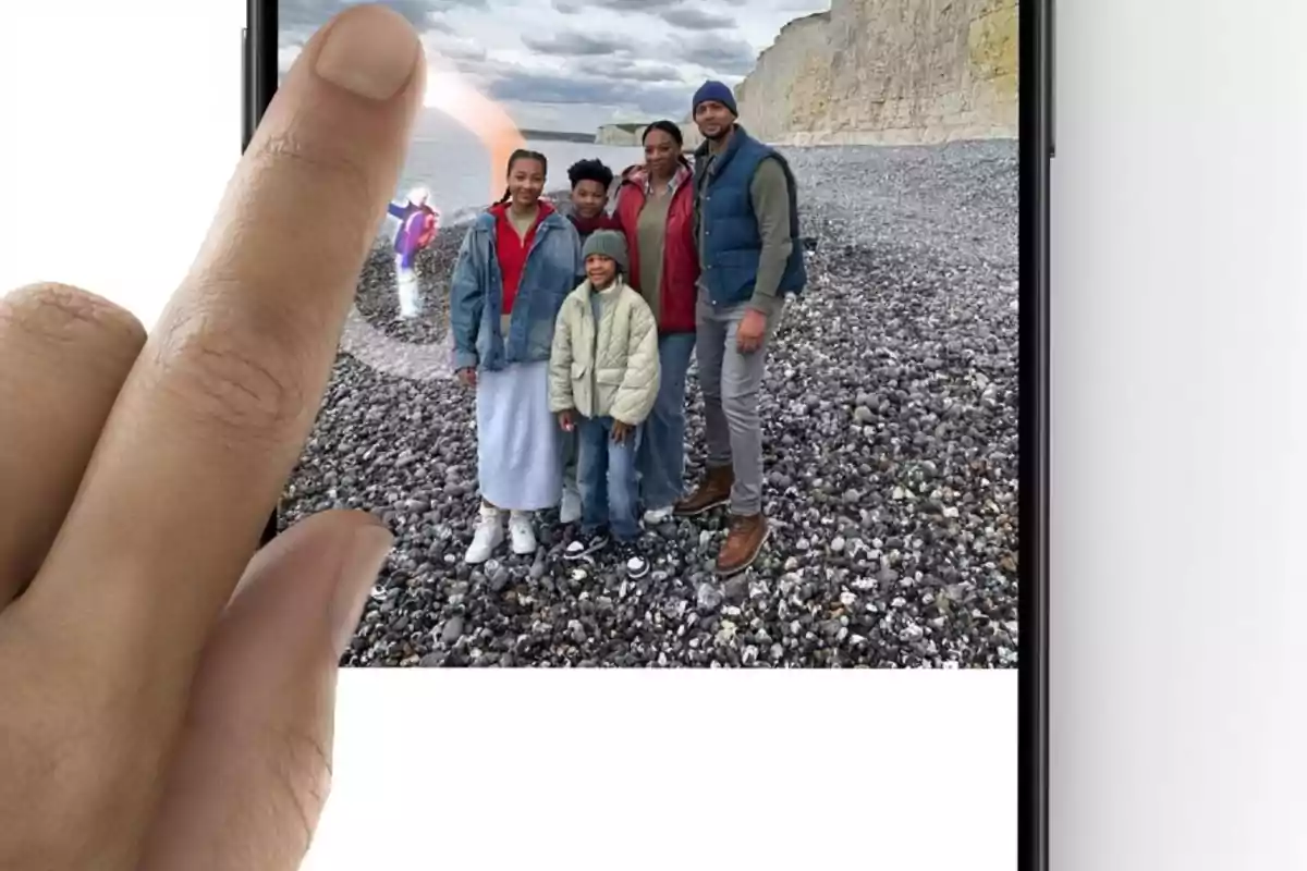 Una mano interactúa con una foto en un teléfono móvil, mostrando a una familia posando en una playa de piedras con acantilados al fondo.