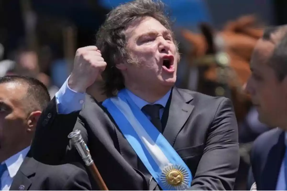 Un hombre con traje y banda presidencial gesticula con entusiasmo en un evento al aire libre.