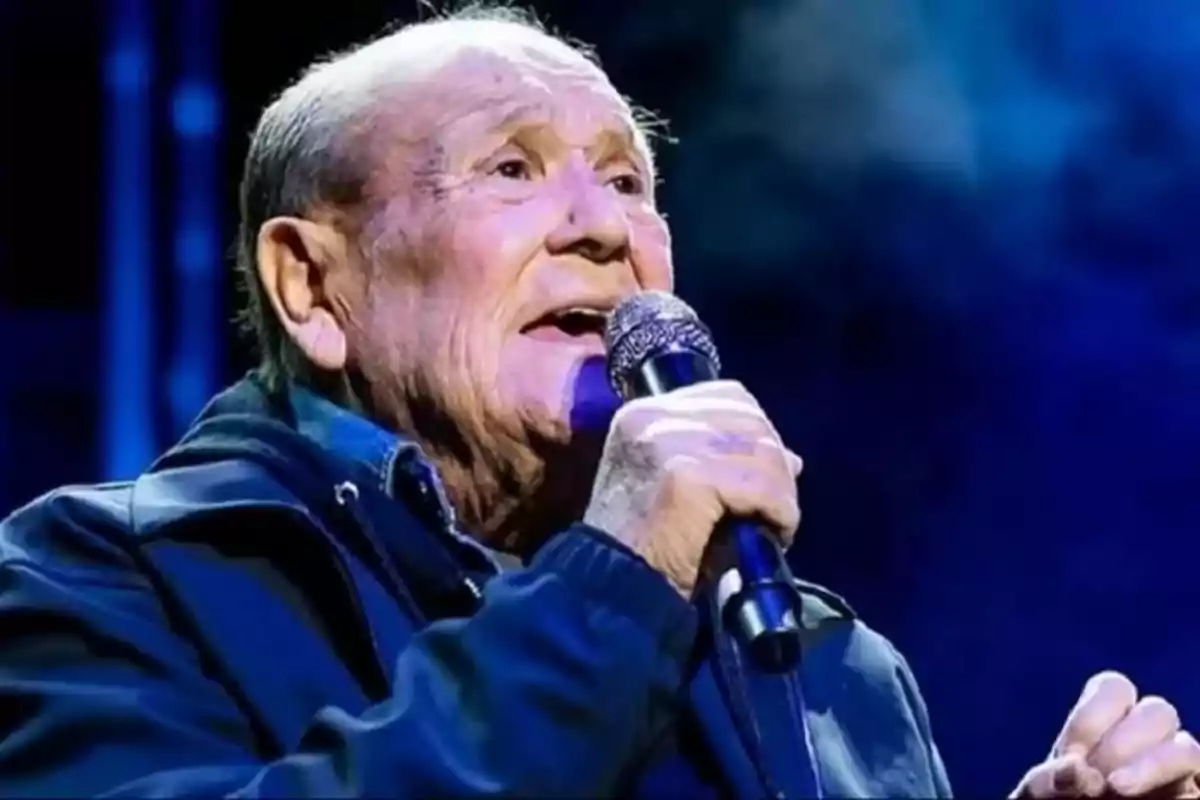 Un hombre mayor cantando en un escenario con un micrófono en la mano y un fondo oscuro.