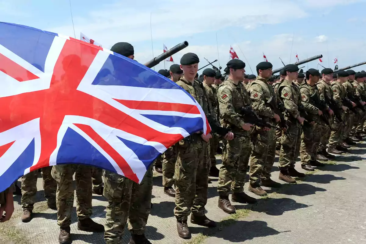 Soldiers in formation with military uniforms and a United Kingdom flag waving in front.