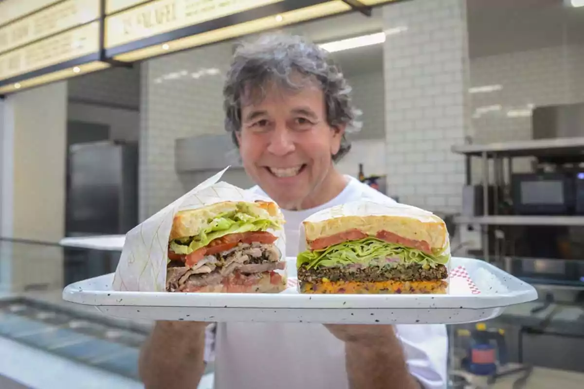 Ariel Rodríguez Palacios sonriendo, sostiene una bandeja con dos mitades de sándwiches grandes y apetitosos en un entorno de cocina moderna.