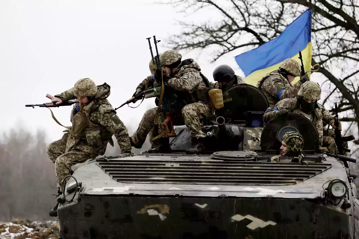 Soldados con uniforme militar camuflado sobre un vehículo blindado con una bandera azul y amarilla ondeando en el fondo.